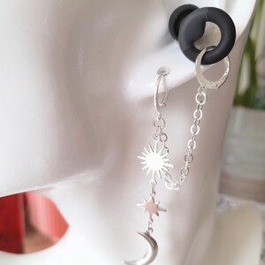 May include: A silver-toned double hoop earring set connected by a delicate chain. One hoop features a dangling charm sequence of a sunburst, a four-pointed star, and a crescent moon. The other hoop is plain, and a black silicone ear gauge is visible.