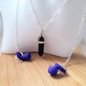 May include: A silver chain necklace with a black crystal pendant and two necklaces with purple earbud pendants. The necklaces are displayed on a white mannequin head and a wooden surface.