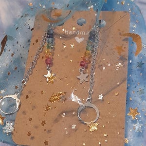 May include: A pair of silver-toned earrings featuring a chain connected to a small handcuff charm and a star charm. The earrings are displayed on a tan card with the text "Handmade" and a heart icon, all inside a blue organza bag with gold stars.