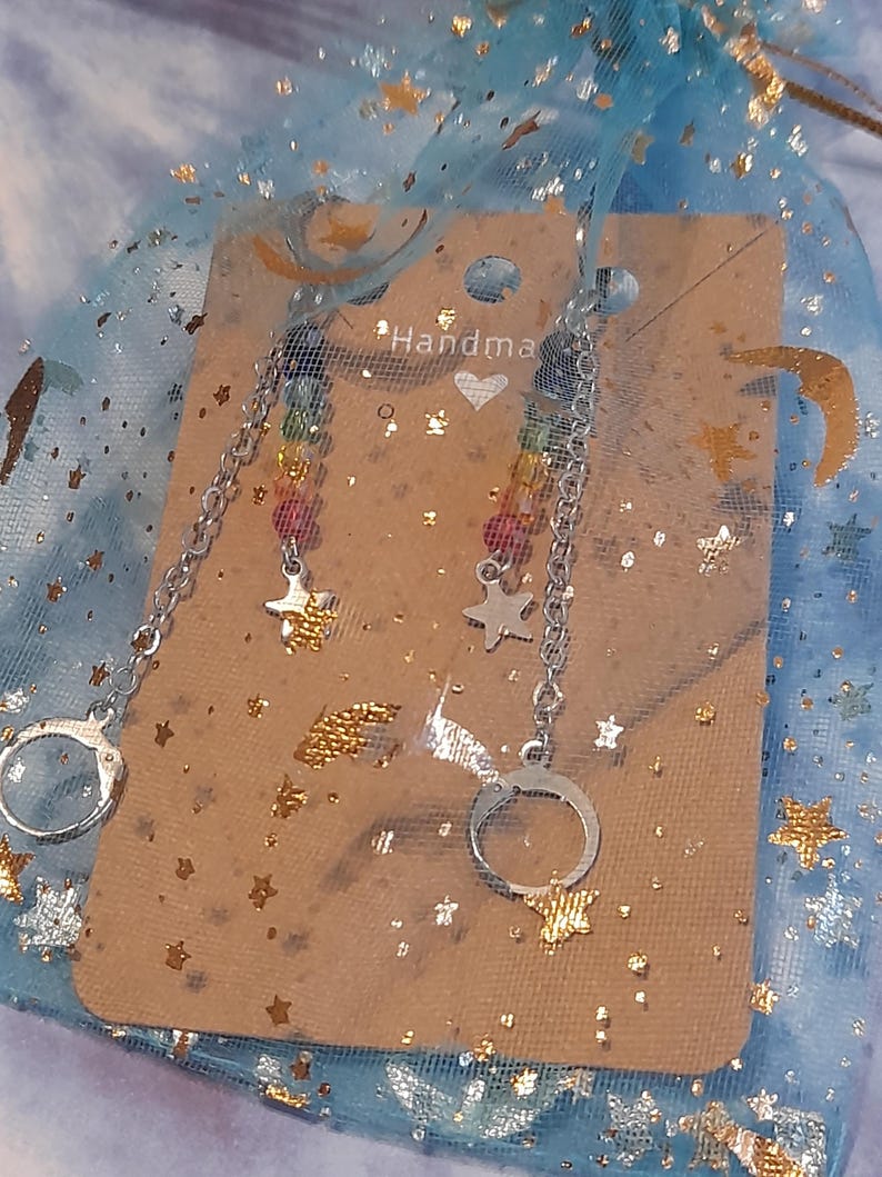 May include: A pair of silver-toned earrings featuring a chain connected to a small handcuff charm and a star charm. The earrings are displayed on a tan card with the text "Handmade" and a heart icon, all inside a blue organza bag with gold stars.