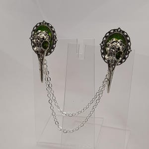 May include: A pair of silver tone bird skull brooches with green enamel accents and a silver chain connecting them.