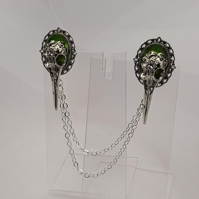 May include: A pair of silver tone bird skull brooches with green enamel accents and a silver chain connecting them.