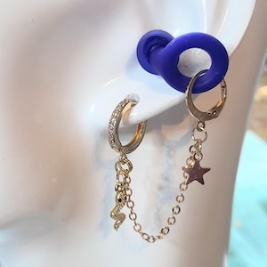 May include: Gold-toned hoop earrings with a dangling chain. One earring features a small snake charm, the other a star. The hoops are adorned with small, clear stones. A blue ear gauge is also visible.