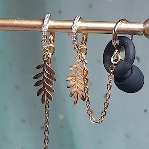 May include: Gold-toned hoop earrings with leaf charms and a chain. One earring has a row of small clear stones. A black earbud is attached to the other earring with a chain. The earrings are displayed on a gold bar.