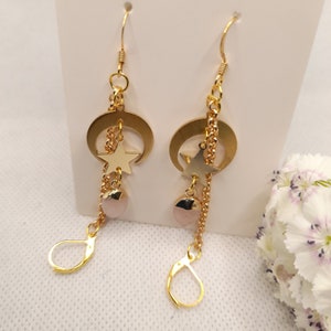 May include: Gold-toned earrings featuring a crescent moon and star design. Each earring has a chain with a star charm, a pink gemstone, and a lever-back closure. The earrings are displayed against a white background.