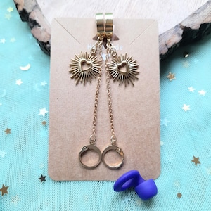 Könnte beinhalten: Goldfarbene Ohrringe mit einem Sonnenstrahlen-Design und herzförmigen Ausschnitten. Die Ohrringe hängen an einer Kette und enden mit einem Handschellen-Design. Am unteren Bildrand befindet sich ein kleines, blaues Silikonobjekt.