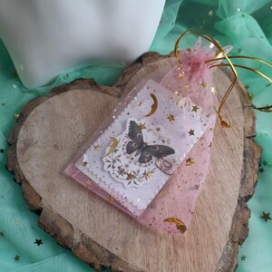 May include: A pink organza gift bag with gold accents holds a small card featuring a butterfly design, crescent moons, and stars. The card is layered over a white card with a scalloped edge. The bag is tied with a gold drawstring, and rests on a wooden heart.