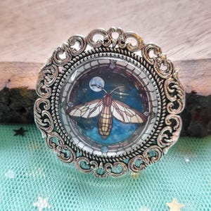 Könnte beinhalten: Runder, silberfarbener Broschenanhänger mit einem verzierten Filigranrand. Die Mitte zeigt ein Design im Stil eines Buntglasfensters mit einer Nachtfalter vor einem blauen Nachthimmel mit Vollmond. Der Nachtfalter hat beige und braune Flügel.