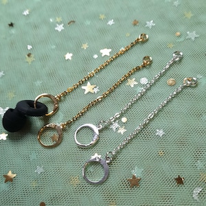 Peut inclure: Chaînes pour écouteurs de couleurs or et argent. Chaque chaîne est dotée d'un petit fermoir à anneau et d'une boucle pour la fixation des écouteurs. Les chaînes sont présentées sur une surface verte avec des confettis en forme d'étoiles.