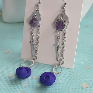 May include: A pair of silver-tone earrings with a unique design. Each earring features a purple gemstone set in a silver oval, a small sword charm, and a chain leading to a purple rose-shaped bead. The earrings hang from a silver hook.
