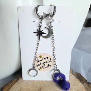 May include: Silver hoop earrings with celestial charms, including a star, crescent moon, and a dark star. The earrings have a chain detail and a purple accent. The text on the card reads "You've got great taste."