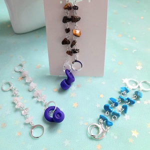May include: An assortment of handmade earrings and keychains. The earrings feature brown and gold stones, while the keychains showcase pink, blue, and turquoise stones. Each piece is connected with silver-colored metal rings and clasps. A purple, ear-shaped charm is also present.