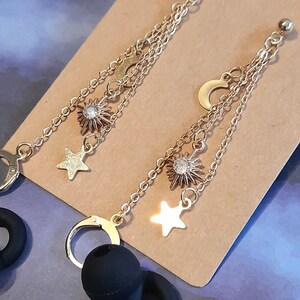 Puede incluir: Pendientes celestiales dorados con un dise&ntilde;o de sol con una gema central, una luna creciente y un charm de estrella. Los pendientes cuelgan de delicadas cadenas y tienen cierre de gancho. Sobre una superficie marr&oacute;n.