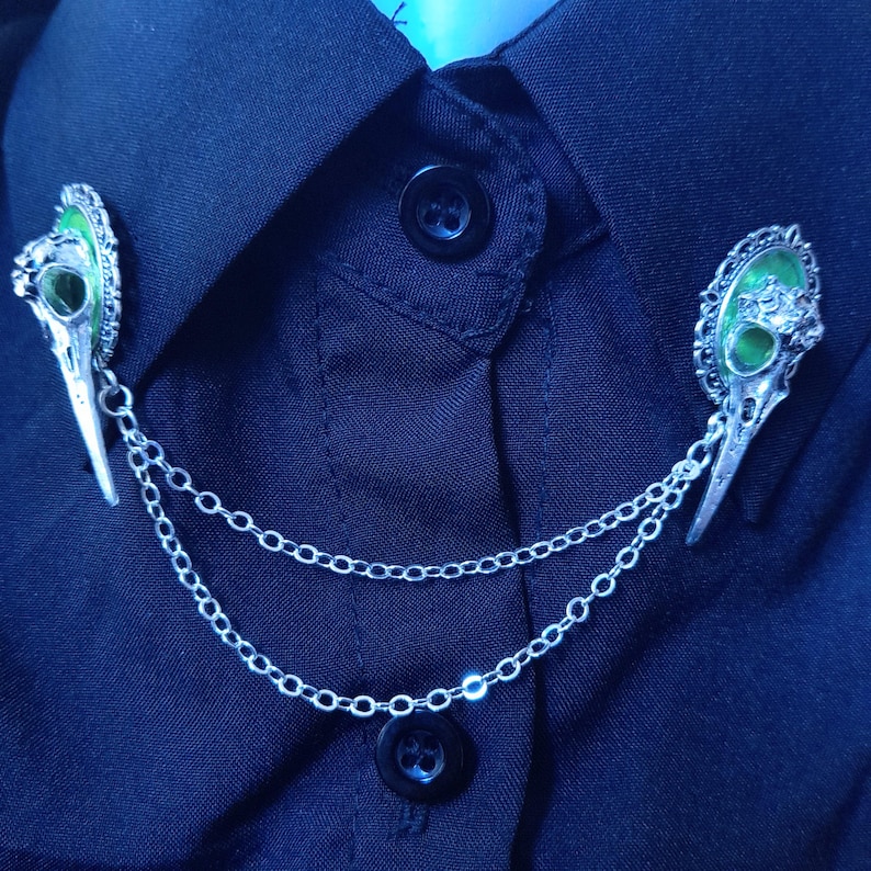 May include: A silver chain collar pin with two green and silver skull charms. The charms are detailed with black eyes and a green enamel finish.