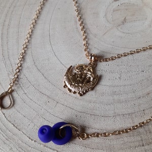 May include: A gold chain necklace with a crescent moon pendant featuring a detailed design. The pendant is attached to a gold chain with a lobster clasp. The necklace is displayed on a wooden surface with a pair of purple earbud tips.