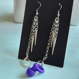 May include: Silver-coloured drop earrings with multiple chains and cone-shaped accents. Each earring features a purple, ear-shaped plug. The earrings are displayed on a black background.