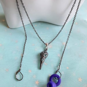 May include: A gunmetal necklace with a bird skull pendant. The skull is intricately detailed and hangs from a fine chain. The necklace is displayed on a white mannequin bust. The background is a light blue.