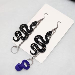 May include: A pair of dangle earrings featuring black snake charms with gold crescent moons and stars on a silver chain. The earrings are displayed on a white background.