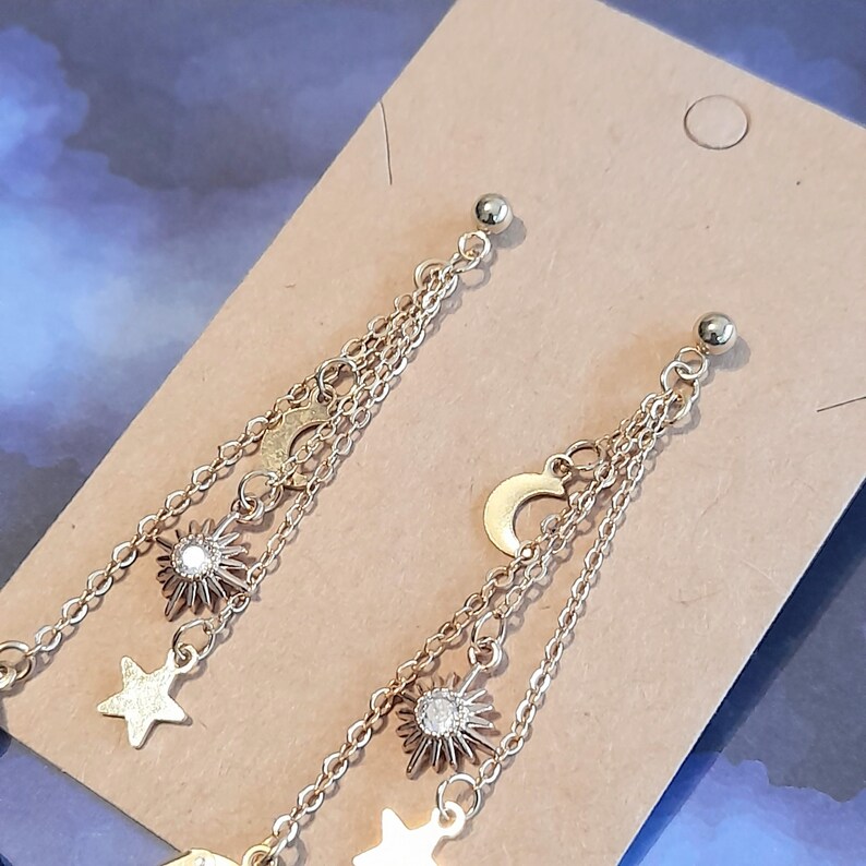 Puede incluir: Pendientes celestiales dorados con colgantes de sol, luna y estrella. Los pendientes tienen m&uacute;ltiples hilos de cadena y un peque&ntilde;o perno redondo en la parte superior. Los colgantes de sol tienen un cristal central. Los pendientes se exhiben en una tarjeta beige.