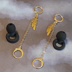 May include: Gold-colored dangle earrings with a feather charm and a small chain. The earrings have a hoop closure and a circular charm at the bottom. Two black earplugs are also in the image.