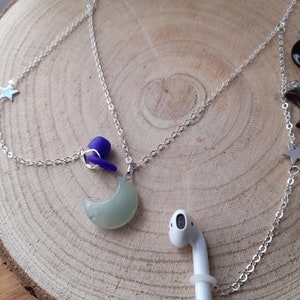 May include: A silver chain necklace with a crescent moon charm, a purple earbud holder, and two star charms. The necklace is on a wooden surface with a white AirPod.