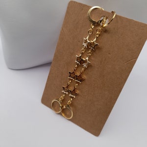 May include: Gold star-shaped earrings with a chain design. The earrings are hanging from a brown card.