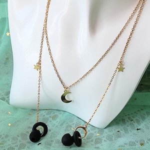 May include: A gold-toned metal chain necklace featuring a central crescent moon charm and two small star charms. The chain extends downwards to connect to two black silicone earbud covers, designed to secure wireless earbuds around the neck.