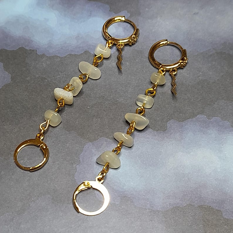 May include: A pair of gold-toned hoop earrings featuring a dangling chain design. Each earring consists of a small circular hoop at the top and bottom, connected by a vertical strand of four smooth, translucent white stones and a small lightning bolt charm.