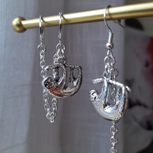May include: Silver-colored sloth earrings. Each earring features a detailed sloth charm hanging from a chain and a hook closure. The sloths are depicted in a hanging position, adding a whimsical touch to the jewelry.
