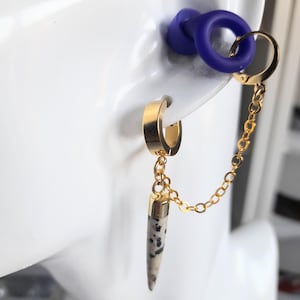 May include: Gold-tone double earrings with a chain. One earring is a small hoop, the other has a chain that connects to a purple ear plug. The dangling earring has a beige and black speckled, cone-shaped charm.