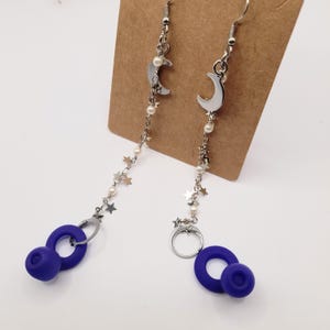 May include: Silver-toned dangle earrings featuring crescent moon charms, small star accents, and faux pearls. The earrings have a chain that leads to a pair of dark blue, circular earbud-shaped charms. The earrings are displayed on a brown card.
