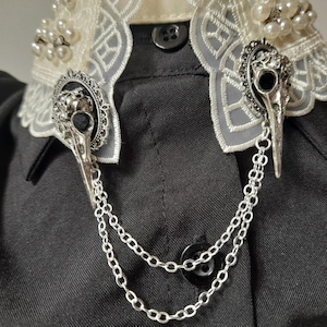 May include: A pair of silver tone skull and raven brooch pins with a silver chain attached. The pins are on a black fabric background.
