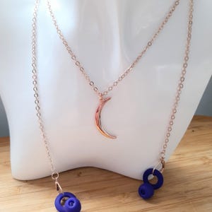 May include: A rose gold necklace with a crescent moon pendant and two strands of chain. The necklace features two blue, circular charms at the ends of the chains. The necklace is displayed on a white mannequin bust.