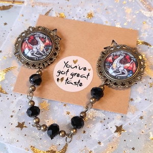May include: A pair of bronze-toned dragon charm necklaces with black beads. The charms feature stained glass-style dragons with red, blue, and yellow accents. The necklaces are connected by a chain and have a tag that reads "You've got great taste".