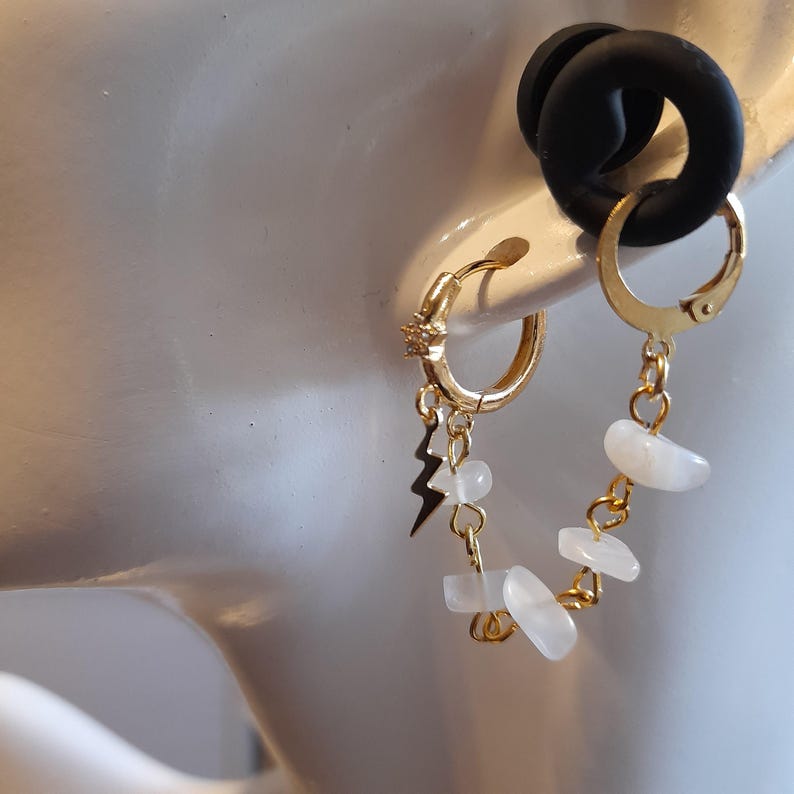 May include: A gold-toned hoop earring featuring a small star charm, a lightning bolt charm, and a delicate chain draped with several small, smooth, white, translucent stone beads. The earring is designed to connect two points on the earlobe.