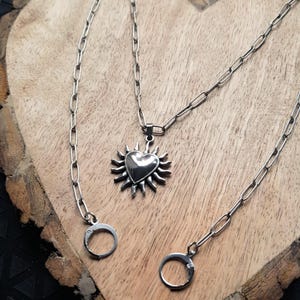Könnte beinhalten: Eine silberfarbene Halskette mit einem herzförmigen Anhänger, der von Sonnenstrahlen umgeben ist. Die Halskette hat eine lange Kette mit einem kreisförmigen Anhänger am Ende. Der Schmuck wird auf einer Holzoberfläche präsentiert.