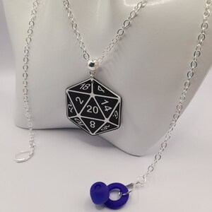 May include: A silver chain necklace with a black 20-sided die pendant. The die has white numbers on it. The necklace also has a pair of blue earbud-style earrings.
