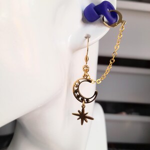 This image shows the loop earplug earings hanging from a model prop of the side of a face and ear. The earring charms hang down from the earhook and the chain with the clip at the end holds a loop earplug. The earplug has been placed into the ear prop to give an example of how the earring will look when the wearer is using the loop earplugs in their ears.