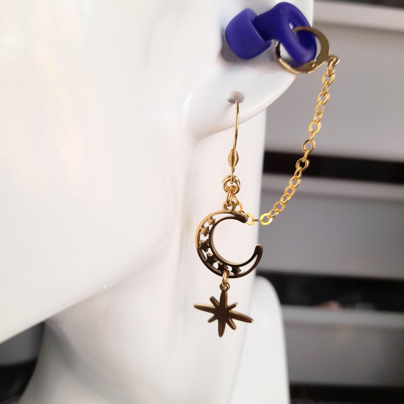 This image shows the loop earplug earings hanging from a model prop of the side of a face and ear. The earring charms hang down from the earhook and the chain with the clip at the end holds a loop earplug. The earplug has been placed into the ear prop to give an example of how the earring will look when the wearer is using the loop earplugs in their ears.