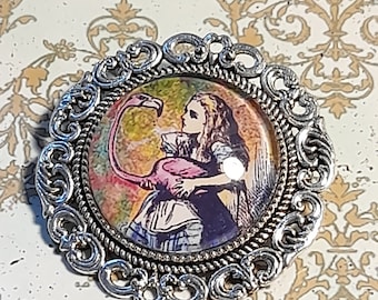 May include: A silver-toned brooch with an ornate design features a colorful image of Alice from Alice in Wonderland holding a pink flamingo.