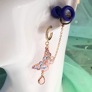 Könnte beinhalten: Goldfarbener Ohrring mit einem Schmetterlingsanhänger und einem kleinen, klaren, tropfenförmigen Anhänger. Der Schmetterling ist mehrfarbig mit rosa, blauen und orangen Farbtönen. Eine kleine Kette verbindet den Ohrring mit einem blauen Ohrstöpsel.