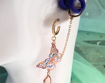 May include: A gold-colored earring with a butterfly pendant and a small, clear teardrop-shaped charm. The butterfly is multi-colored with pink, blue, and orange hues. A small chain connects the earring to a blue ear plug.