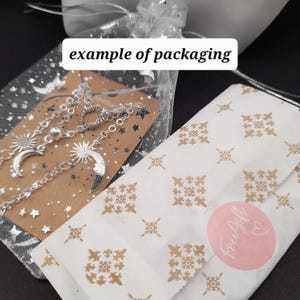 May include: Example of packaging for a silver necklace with a moon and sun charm. The necklace is packaged in a brown paper bag with silver stars and a white tulle bag. The package also includes a pink sticker that says "Free Gift".