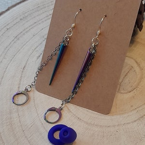 May include: A pair of iridescent earrings with a spike and chain design. The earrings are gold-toned and have a dangling chain with a spike at the end. The spikes are iridescent and have a purple, blue, and green colour.