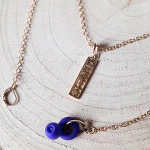 May include: A gold necklace with a pendant featuring a series of cut-out symbols. The pendant is attached to a delicate gold chain. The necklace is displayed on a light wood surface with a purple earbud.