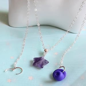 May include: Three delicate silver necklaces with different pendants. One features a star-shaped amethyst, another a purple flower, and the third a silver clasp. The necklaces are displayed on a light blue surface.