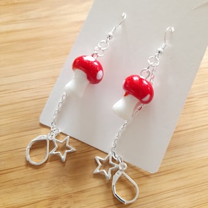 May include: A pair of quirky earrings with red mushroom charms, white spots, and stems. The earrings hang from silver-coloured hooks and chains, with a star-shaped charm and a lever back closure. The earrings are displayed on a white card.