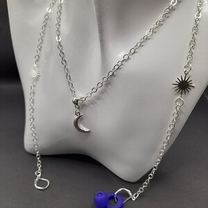 May include: Silver chain necklace with a crescent moon charm and two sun charms. The necklace is on a white mannequin bust. A blue silicone earplug is in the foreground.