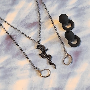 May include: A black metal necklace featuring a sword and snake pendant, a chain with a gold-coloured clasp, and two black earbud tips. The jewellery is set against a light blue and white marbled background.