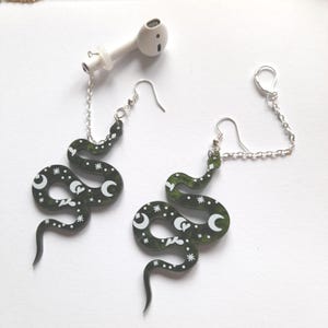 May include: A pair of dark green snake earrings with white crescent moons, stars, and heart designs. The earrings are connected to a silver chain and ear hooks. An earbud is attached to one earring.